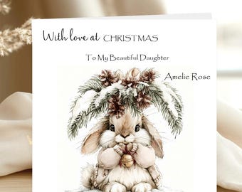 May include: A square greeting card featuring a watercolor illustration of a fluffy brown rabbit holding a gold bell under snow-covered pine branches. The text reads: With love at CHRISTMAS, To My Beautiful Daughter, and Amelie Rose.