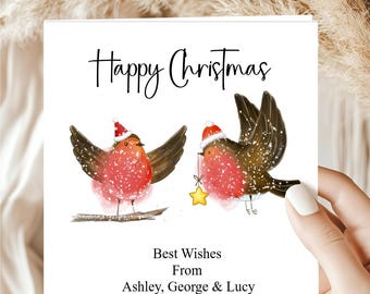 May include: A square white greeting card featuring two illustrated robins wearing red Santa hats. One robin sits on a branch, the other flies holding a yellow star. Text reads Happy Christmas and Best Wishes From Ashley, George & Lucy.