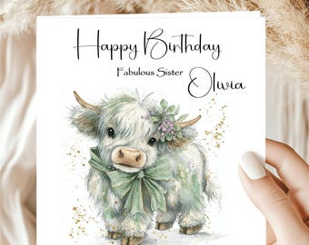 May include: A birthday card featuring a watercolor illustration of a Highland cow with a green bow and floral accents. The card reads "Happy Birthday Fabulous Sister Olivia" in elegant script, with gold speckles.