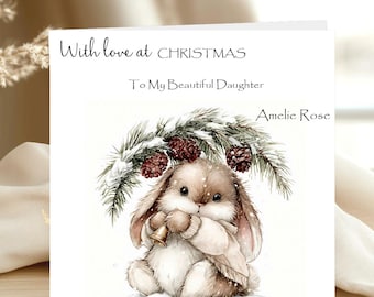 May include: A square greeting card featuring the text With love at CHRISTMAS, To My Beautiful Daughter, and Amelie Rose. The illustration shows a brown bunny wearing a beige sweater, holding a small bell under snow-covered pine branches.