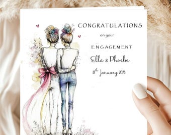 May include: A white engagement card held by a hand, featuring an illustration of two women embracing. The card reads "CONGRATULATIONS on your ENGAGEMENT Ella & Phoebe 12th January 2026". The illustration is in watercolor style with a pink bow.