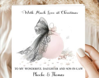 May include: A square Christmas greeting card featuring a pale pink bauble adorned with a large, intricate black lace bow. The text reads With Much Love at Christmas, TO MY WONDERFUL DAUGHTER AND SON-IN-LAW, and the names Phoebe & Thomas.