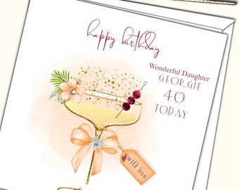 Könnte beinhalten: Eine weiße Karte mit einer rosa und goldenen Illustration eines Cocktailglases mit einer Kirschengarnierung. Der Text "happy birthday" ist in rosa Schrift geschrieben. Der Text "Wonderful Daughter Georgie 40 Today" ist in Burgunderrot geschrieben. Der Text "with love" ist auf einem kleinen braunen Etikett geschrieben.