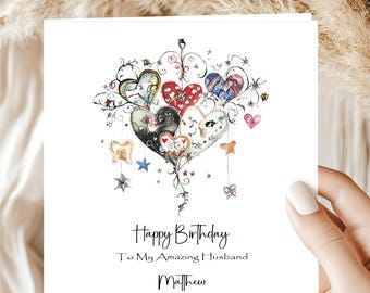 May include: A white greeting card with a watercolor design of colorful hearts and floral accents. The card reads "Happy Birthday To My Amazing Husband Matthew" in black script. The card is held by a person.