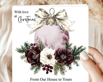 May include: A square greeting card featuring a watercolor illustration of a pale pink Christmas ornament topped with a large cream-colored bow. Below the ornament are dark red flowers, a white flower, pine needles, and berries. Text reads: With love at Christmas, From Our House to Yours.
