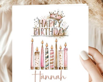 May include: A square greeting card featuring a watercolor illustration of eight lit candles in shades of pink, gold, and white. Above the candles, the text reads Happy Birthday in a decorative font with a pink crown. The name Hannah is printed below.