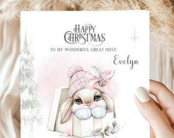 May include: A square Christmas greeting card featuring a white gift box with a cute bunny wearing a pink knit hat and bow. The text reads Happy Christmas To My Wonderful Great Niece Evelyn. A silver pine tree and a star are printed on the card.