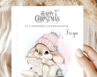 May include: A square greeting card featuring a light brown bunny wearing a pink knit hat with a pom-pom. The text reads Happy 1st Christmas to a wonderful granddaughter Freya. The bunny holds a scroll with a red seal and sits by a silver bell.