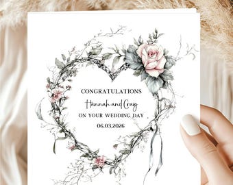 May include: A white wedding card with a heart-shaped floral wreath design. The wreath features pink roses, gray leaves, and delicate branches. The card reads "Congratulations Hannah and Craig on your wedding day 06.03.2026."