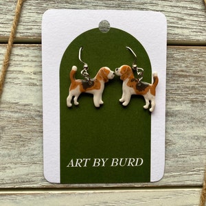 May include: A pair of dangle earrings featuring a brown and white beagle dog design. The earrings are on a white card with a green background and the text "ART BY BURD".
