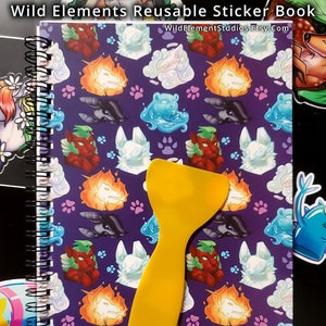 May include: A reusable sticker book with a purple cover featuring various fantasy creature designs and paw prints. A yellow applicator tool rests on the cover. The text "Wild Elements Reusable Sticker Book" is at the top.
