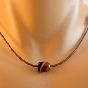 May include: A brown leather necklace with a central, barrel-shaped bead. The bead has brown, black, and white stripes. The necklace is displayed on a person's neck.