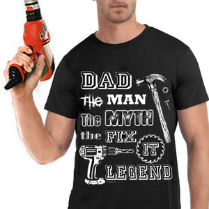 Puede incluir: Camiseta negra con un gráfico blanco que dice "Dad The Man The Myth The Fix It Legend" con imágenes de herramientas como un martillo, un destornillador y una sierra.