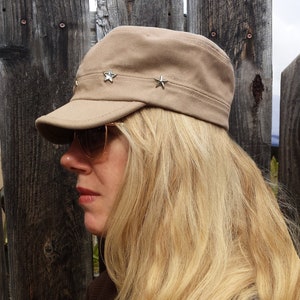 May include: A woman with long blonde hair is wearing a brown cap with silver star embellishments. The cap has a curved brim and a structured crown. She is wearing a brown sweater.