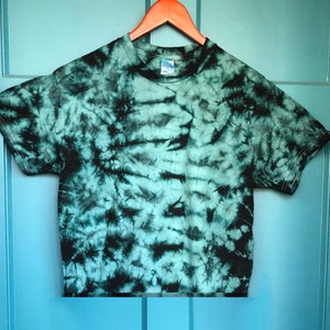 Puede incluir: Una camiseta de manga corta con un estampado tie-dye en tonos turquesa y negro cuelga de una percha naranja. La camiseta se muestra contra una puerta azul claro, creando un fondo contrastante. El diseño tie-dye presenta un patrón único y arremolinado.