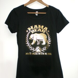 Puede incluir: Camiseta negra con un dise&ntilde;o de oso en oro en relieve dentro de una corona con el texto "Mama Bear" y "Don't Mess With Mama".