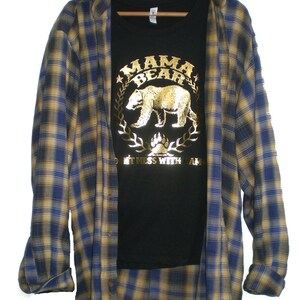 Puede incluir: Una camiseta negra con un dise&ntilde;o de oso en l&aacute;mina dorada y el texto "Mama Bear Don't Mess With Mama". La camiseta est&aacute; superpuesta a una camisa de franela a cuadros azules y amarillos.