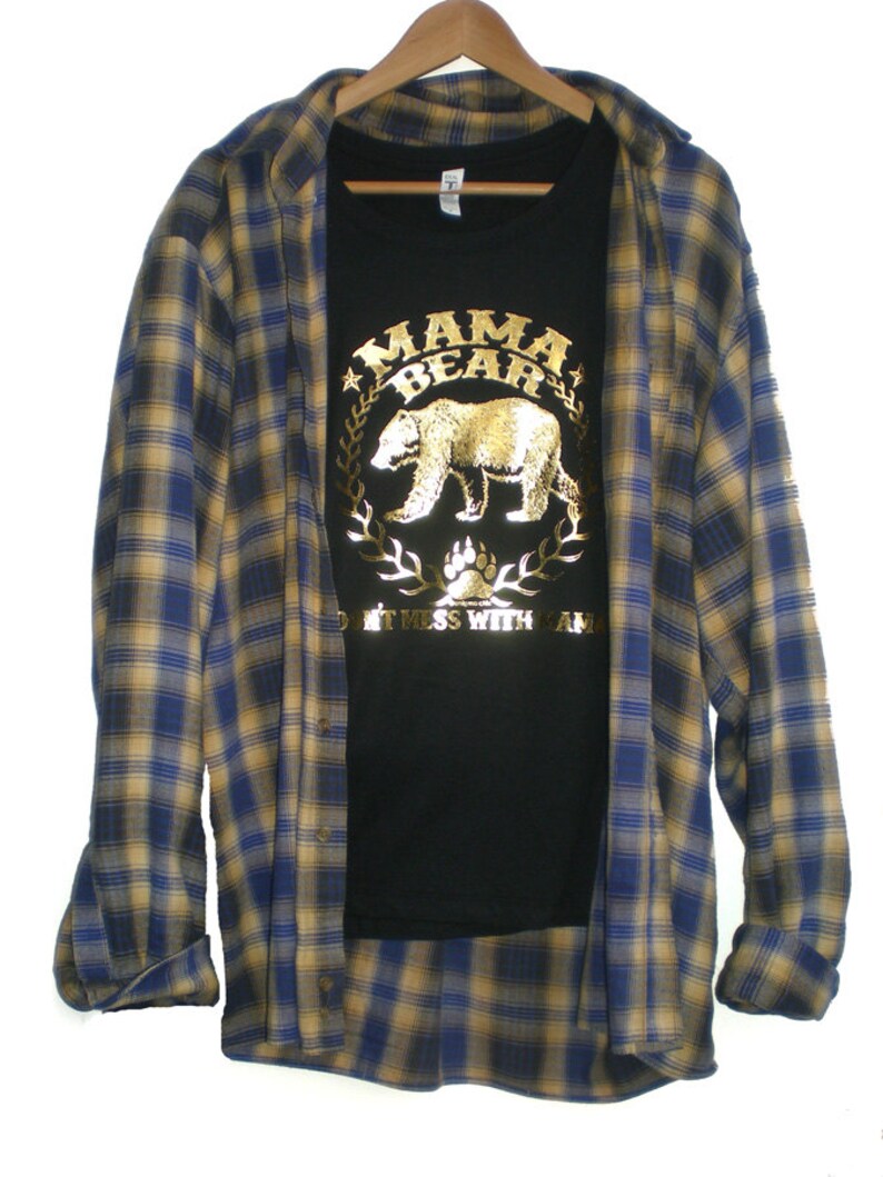 Puede incluir: Una camiseta negra con un dise&ntilde;o de oso en l&aacute;mina dorada y el texto "Mama Bear Don't Mess With Mama". La camiseta est&aacute; superpuesta a una camisa de franela a cuadros azules y amarillos.