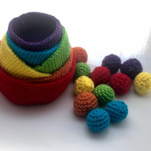 Puede incluir: Un conjunto de cuencos anidados de crochet de colores arcoíris con un conjunto de bolas de crochet de colores. Los cuencos están apilados en orden de color, desde el morado hasta el rojo. Las bolas son de varios colores, incluyendo morado, rojo, naranja, amarillo, verde y azul.