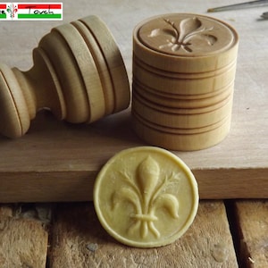 May include: Two wooden cookie stamps with a fleur-de-lis design. The stamps are made of light wood and have a natural finish. A cookie with the fleur-de-lis design is also shown.