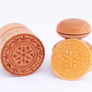 Op de afbeelding: Twee houten koekjesvormen met ingewikkelde gesneden ontwerpen. Een vorm is een cilinder met een cirkelvormig ontwerp, de andere is een ronde vorm met een verhoogd handvat. De ronde vorm wordt getoond met een gele koekjesafdruk.