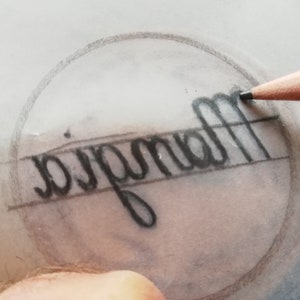 May include: A close-up of a pencil drawing on a piece of tracing paper. The drawing is of the word "Magnolia" inside a circle.