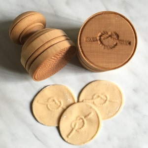 Op de afbeelding: Een houten koekjesvormpje met een touwknots-ontwerp. De vormpje wordt gebruikt om decoratieve koekjes te maken. Drie koekjes met het touwknots-ontwerp zijn te zien.