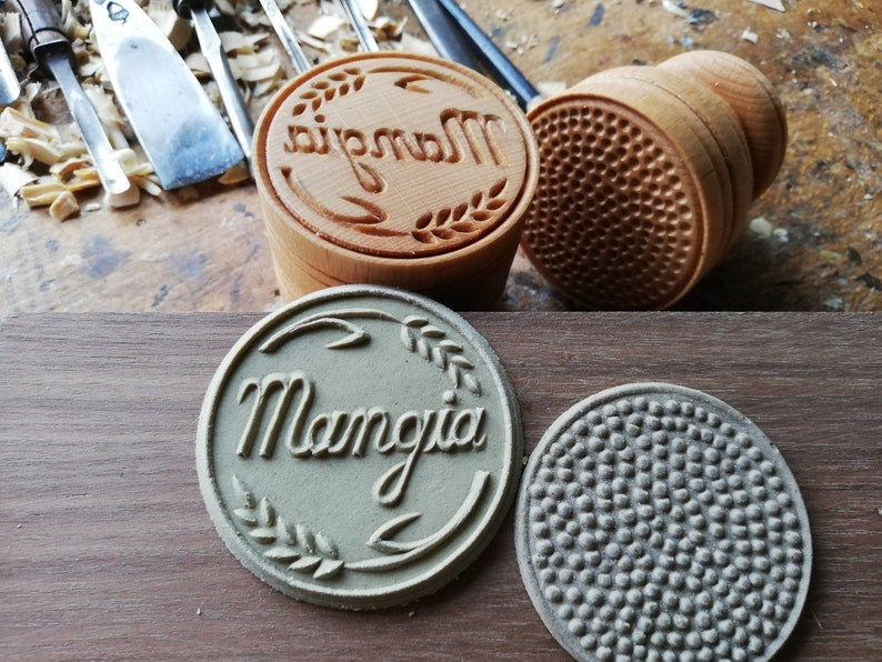 May include: Two wooden dough stamps, one with the word "Mangia" carved into it and a wheat design, and the other with a textured pattern. The stamps are on a wooden surface.