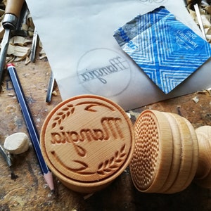 May include: Two wooden stamps, one with the word "Mangia" carved into it and a wheat design, and the other with a textured pattern. The stamps are on a wooden surface with wood shavings.