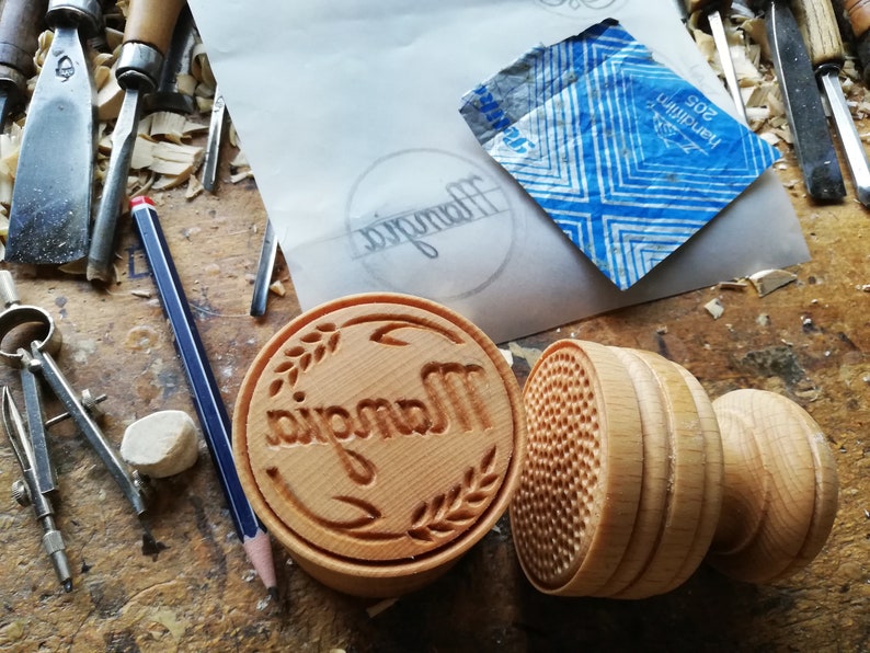 May include: Two wooden stamps, one with the word "Mangia" carved into it and a wheat design, and the other with a textured pattern. The stamps are on a wooden surface with wood shavings.