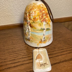 May include: A ceramic bell with a painted autumn scene of trees and a river. The bell is attached to a brown cord and has a small ceramic tag with the word "Stefani" written on it.
