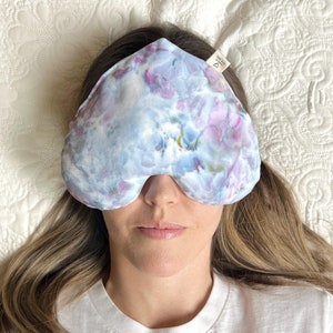 May include: A blue and purple tie-dye eye mask in a heart shape. The mask is lying on a white bedspread.