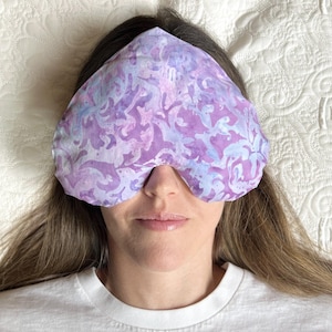 Könnte beinhalten: Eine herzförmige Augenmaske mit einem violetten und blauen Muster. Die Maske liegt auf einem weißen Bett.