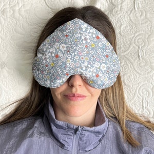 May include: A person wearing a grey floral eye mask while lying down on a white bed.