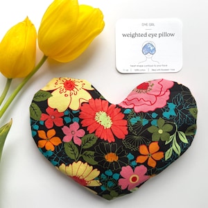 May include: A heart-shaped weighted eye pillow with a floral pattern in red, pink, yellow, and orange on a black background. The pillow is next to two yellow tulips and a card that reads "weighted eye pillow".
