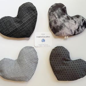 May include: Four heart-shaped weighted eye pillows in various patterns: black and white grid, gray tie-dye, gray and white stripes, and gray with black polka dots. A card reads "weighted eye pillow".