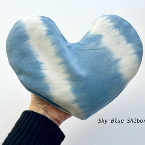 Puede incluir: Una almohada con forma de coraz&oacute;n con un patr&oacute;n de tie-dye azul cielo y blanco. La almohada est&aacute; hecha de tela y tiene una superficie suave y texturizada. "Sky Blue Shibori" est&aacute; escrito en blanco debajo de la almohada.