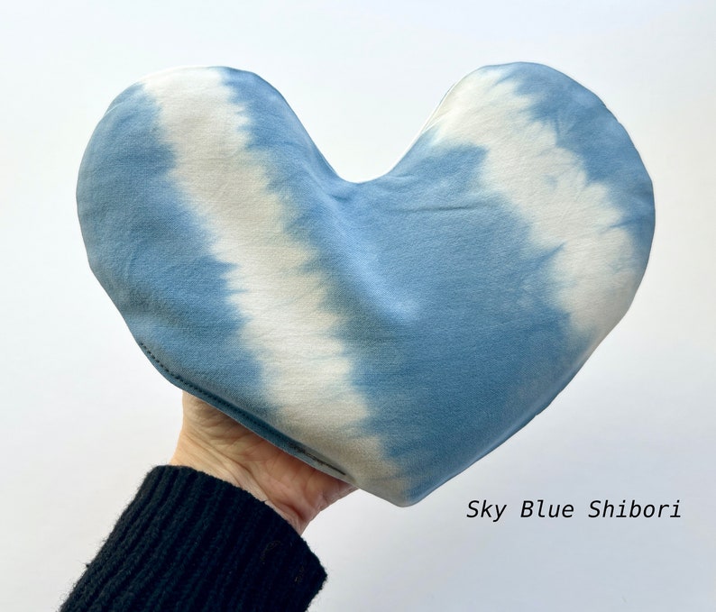 Puede incluir: Una almohada con forma de coraz&oacute;n con un patr&oacute;n de tie-dye azul cielo y blanco. La almohada est&aacute; hecha de tela y tiene una superficie suave y texturizada. "Sky Blue Shibori" est&aacute; escrito en blanco debajo de la almohada.
