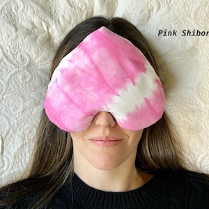 Puede incluir: Una m&aacute;scara para dormir de color rosa y blanco con el texto "Pink Shibori" en la esquina superior derecha. La m&aacute;scara la lleva puesta una persona que est&aacute; tumbada sobre una almohada blanca.