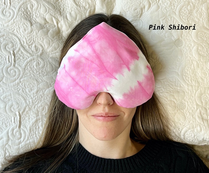Puede incluir: Una m&aacute;scara para dormir de color rosa y blanco con el texto "Pink Shibori" en la esquina superior derecha. La m&aacute;scara la lleva puesta una persona que est&aacute; tumbada sobre una almohada blanca.