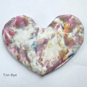 Puede incluir: Una almohada de tela tie-dye en forma de coraz&oacute;n con fondo blanco y remolinos rosas, azules y amarillos.