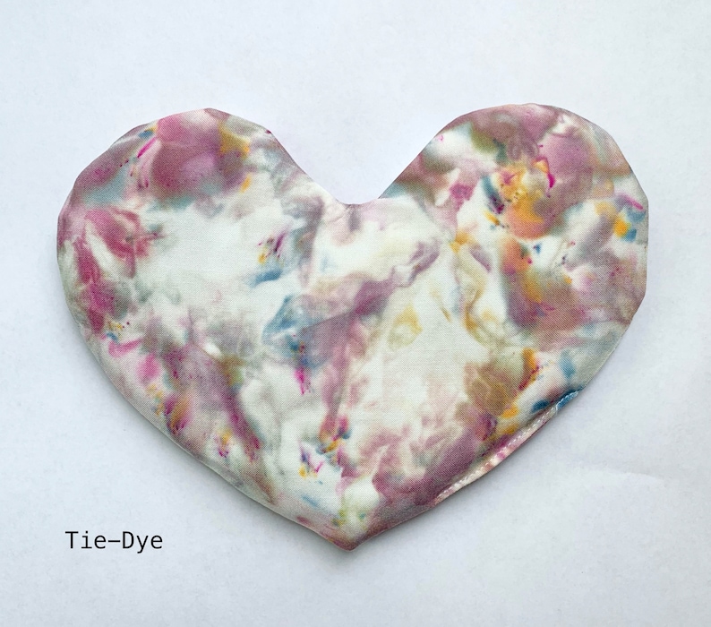 Puede incluir: Una almohada de tela tie-dye en forma de coraz&oacute;n con fondo blanco y remolinos rosas, azules y amarillos.