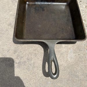 Puede incluir: Una sartén cuadrada de hierro fundido con una superficie oscura y curada, que cuenta con un mango único con una abertura ovalada alargada. La sartén tiene lados rectos y verticales y una base plana, con un acabado metálico gris oscuro y negro.