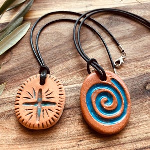 Pode incluir: Dois colares com pingentes de cerâmica feitos à mão. Um pingente apresenta um desenho de cruz com detalhes turquesa, o outro uma espiral azul. Ambos estão em cordões pretos. Os pingentes são de uma cor terracota quente.