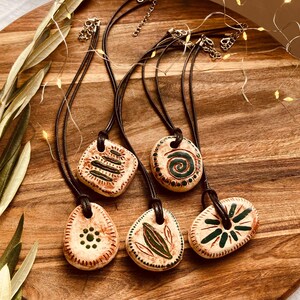 May include: Five ceramic pendant necklaces with brown leather cords. Each pendant has a unique green and brown design, including geometric patterns and leaf motifs. The necklaces are displayed on a wooden surface.