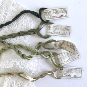 May include: Five twisted skeins of embroidery thread in varying shades of green, ranging from dark forest to light olive. Each skein features a white paper label printed with washing instructions and shade numbers 369, 333, 002, 332, and 331.