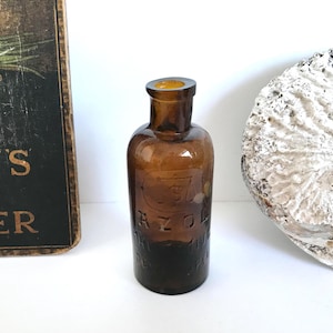 May include: A small, antique brown glass bottle with a rounded body and a short neck. The bottle has embossed lettering and a dark amber color. The bottle is likely a vintage apothecary or medicine bottle.