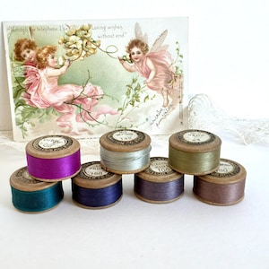 May include: Seven wooden spools of thread in various colours including purple, teal, silver, sage green, dark blue, and brown, arranged in front of a vintage postcard featuring two winged figures and the text Through the telephone I send Loving wishes without end.