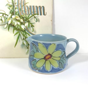 May include: A light blue ceramic creamer with a large yellow and green floral design. The creamer has a handle and a spout. The word "Album" is visible in the background.
