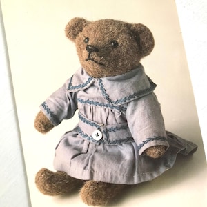 May include: A handmade brown teddy bear wearing a light blue dress with a Peter Pan collar and embroidered details. The dress has a button closure. The bear is sitting on a white surface.